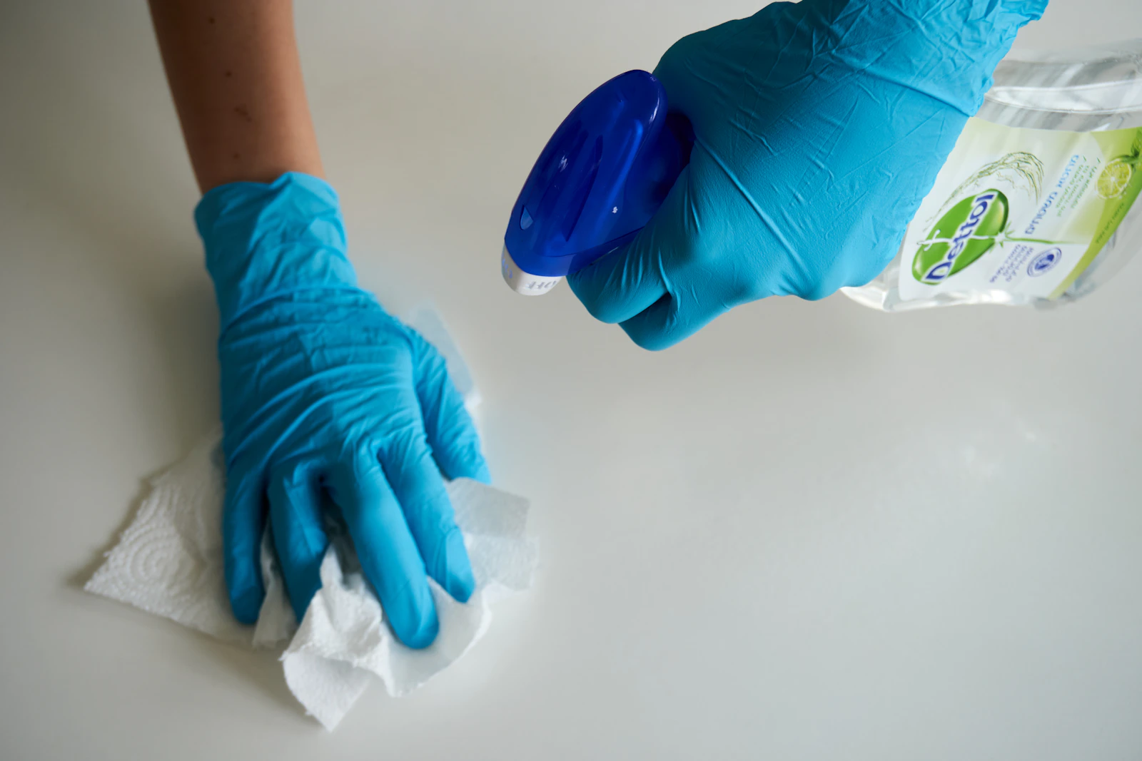 Blue-gloved hands using a spray bottle and cloth to clean a white surface